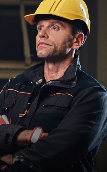 A man wearing a yellow hard hat and a dark work jacket with orange trim looks to the side.